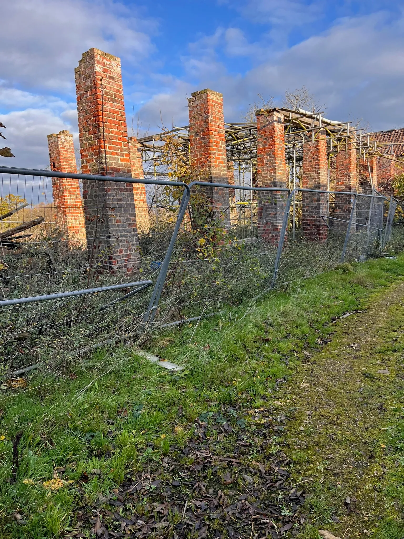 Crumbling red brick pillars behind temporary fencing and scaffolding, surrounded by overgrown grass