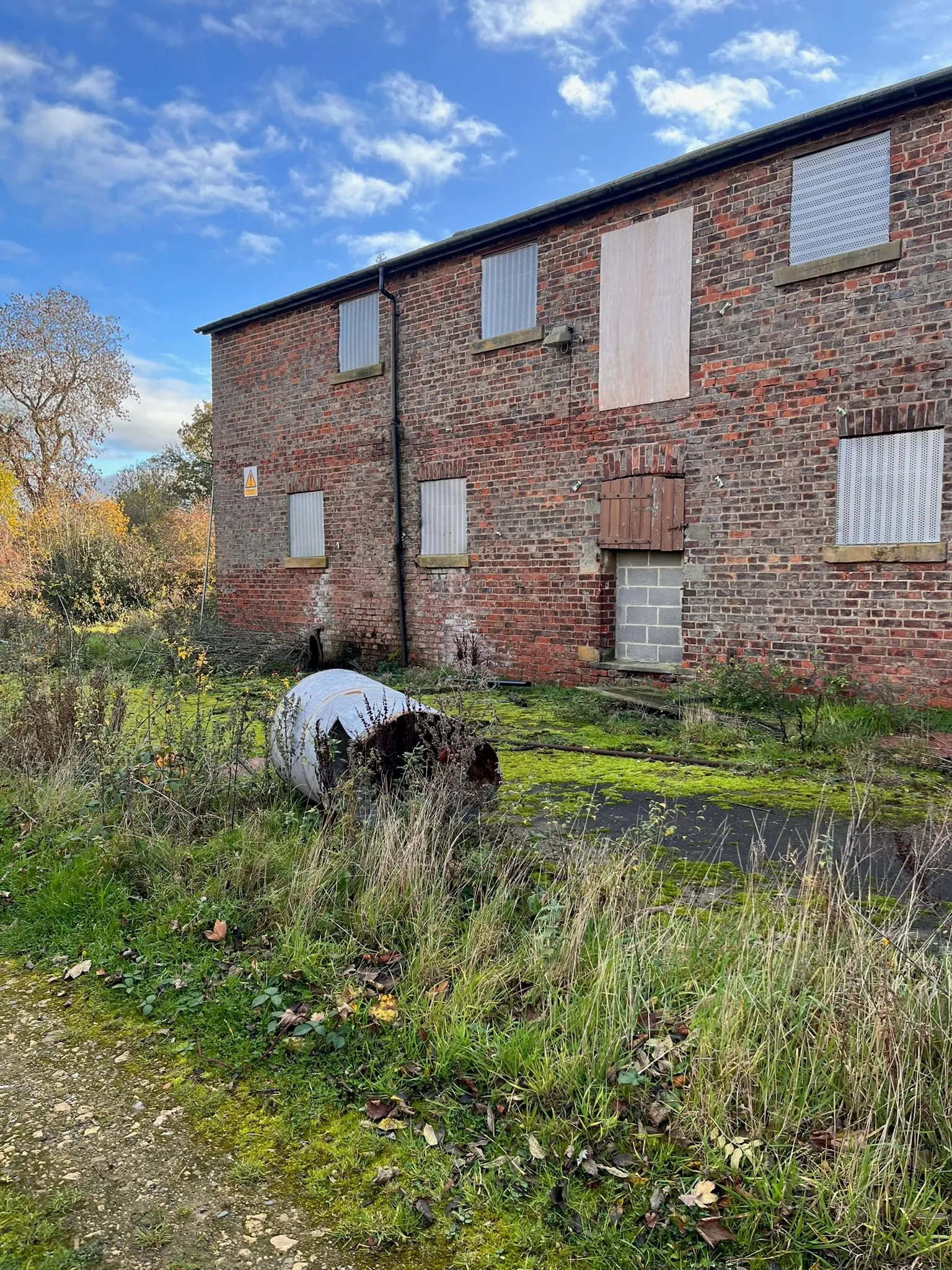A two-storey brick farm building with its windows boarded up, beside an overgrown, moss-covered yard