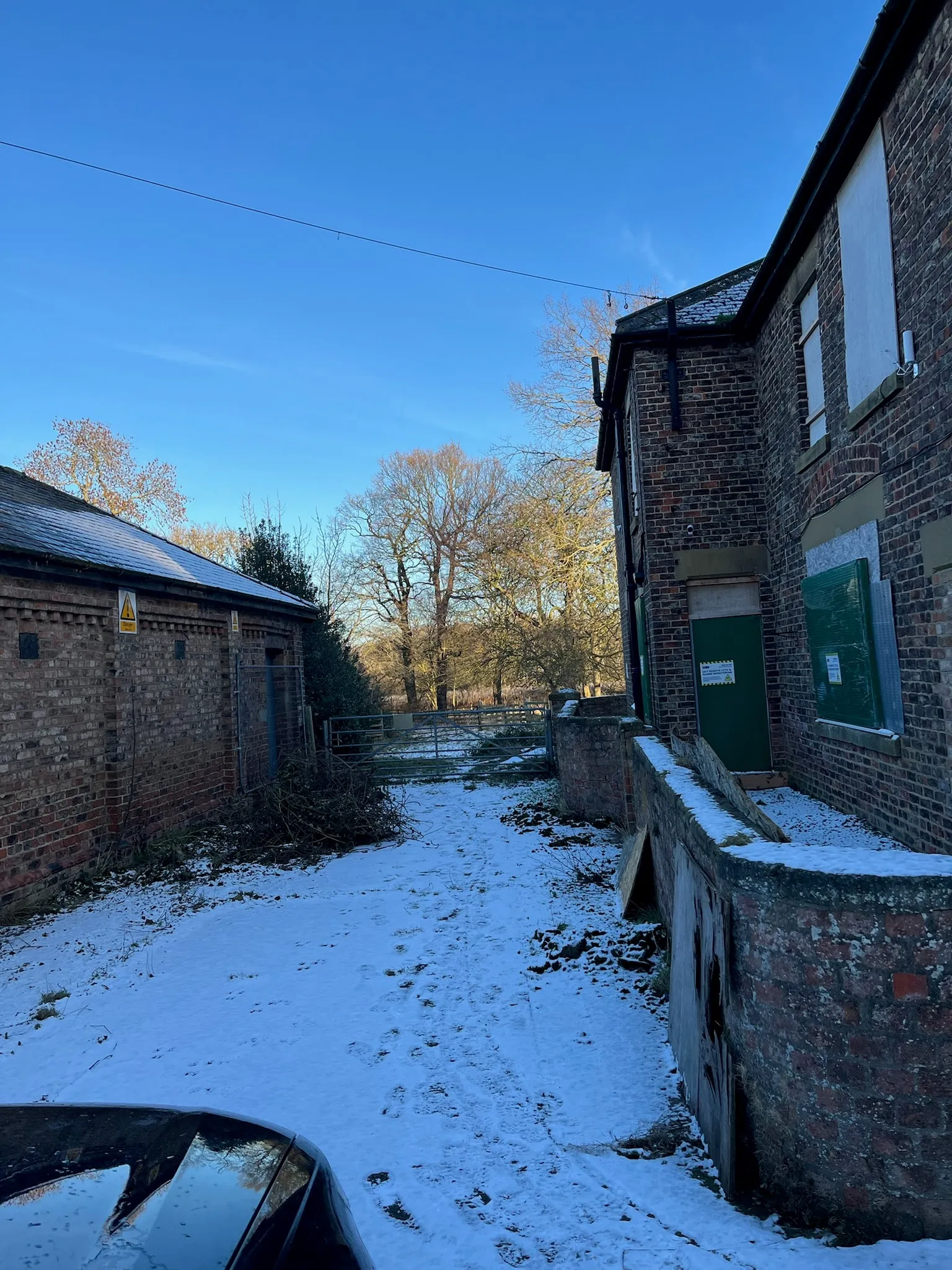 Snow in the yard between the boarded-up farmhouse and a brick outbuilding
